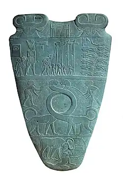 Reverse of the Narmer Palette, circa 3100 BC. The top row depicts four men carrying standards. Directly above them is a serekh containing the name of the king, Narmer.