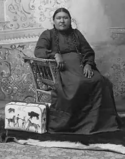 A black and white photograph of Nellie Two Bears Gates. She is sitting in a chair with a beaded valise at her feet.