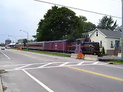 A train parked along a railroad track, in front of and parallel to a road. The train includes a purple and black diesel locomotive numbered 6, a baggage car, and several passenger cars labeled "Newport Dinner Train".