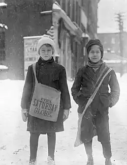 Two young newsboys smiling and standing in the snow. One boy is holding a bag.