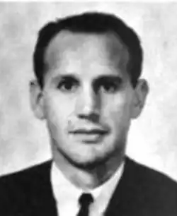 A young man with dark hair and eyes, he has a receding hairline, is cleanshaven, and wearing a coat and tie