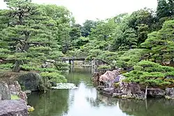 A pond with stones along the sides located in a garden with pine and other trees.