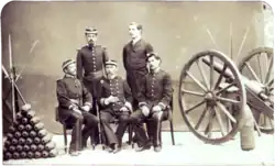 A photograph depicting a group of five uniformed men posed between a pyramid of artillery shells on the left and a wheeled field artillery piece on the right