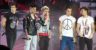 Five young adult males standing next to each other on a stage in Glasgow, Scotland, with microphones in their hands. Member Niall Horan is the only one visibly singing. From left to right: Harry Styles, Zayn Malik, Niall Horan, Liam Payne, and Louis Tomlinson.