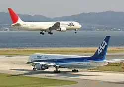 A white and red-tailed Japan Airlines aircraft above the runway, with landing gears down, and an All Nippon Airways in blue and white livery taxiing
