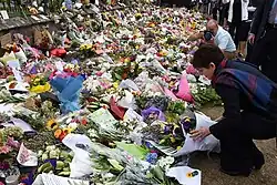 A woman adds a flower arrangement to a large memorial display set against a fence.