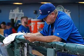 A man in a blue shirt and cap leans on a dugout railing looking over a lineup card.