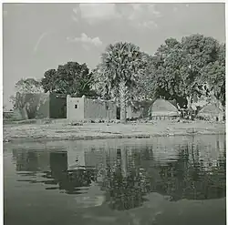 Black-and-white photo of small mudbrick houses and thatched huts next to a river.