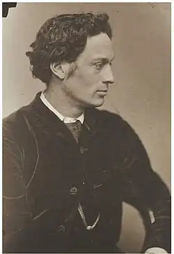 A half-length black and white photograph of a man in profile. He is clean-shaven, with medium-length wavy hair. He wears a dark, velvety suit jacket.