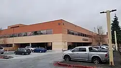 Isometric photo of the building showing the south (entrance) and west sides. It is a two story flat topped building. The walls are horizontally banded in colors; tan on the bottom, darker tan in the middle, and terra cotta red on the top. The south side of the building appears more faded than the other side.