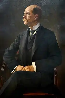 George is pictured as a balding man in his sixties with a large handlebar moustache, wearing a three-piece suit, seated in a chair