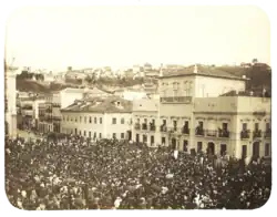 An old photograph showing a crowded square in front of a large, white, multi-storied building