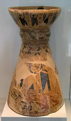 Photograph of a complete stand: a vaguely hourglass-shaped ceramic object, its base much longer than its top, with painted decoration. Most is badly damaged, but the figures of bearded men can be seen.