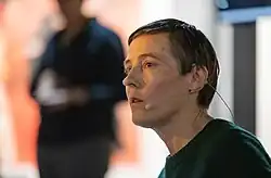 3/4 headshot of Lisa with headphone mic. She has short hair. An out-of-focus figure is in the background.