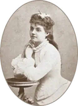 teenage white woman in 19th-century costume seated, with both elbows on a side table and her hands to her face