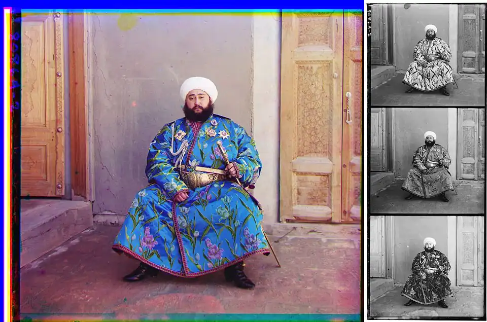 A large color photograph abutting (to its right) a column of three stacked black-and-white versions of the same picture. Each of the three smaller black-and-white photos are slightly different, due to the effect of the color filter used. Each of the four photographs differs only in color and depict a turbaned and bearded man, sitting in the corner an empty room, with an open door to his right and a closed door to his left. The man is wearing an ornate full-length blue robe trimmed with a checkered red-and-black ribbon. The blue fabric is festooned with depictions of stems of white, purple, and blue flowers. He wears an ornate gold belt, and in his left hand, he holds a gold sword and scabbard. Under his right shoulder strap is a white aiguillette; attached to his robe across his upper chest are four multi-pointed badges of various shapes, perhaps military or royal decorations.