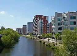 The canalised River Aire flows from the Dark Arches under Leeds's main railway station towards the bottom of the picture. To the left of the river is the lock which links the river with the Leeds and Liverpool Canal. To the right is a riverside walk beneath modern buildings, and in the distance, beyond the railway viaduct and station, are more high-rise modern buildings located on the west side of the city centre.