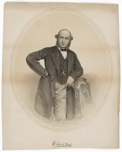 A lithograph with only grey and cream colours. It features a man wearing a three-piece suit and bow tie leaning to his left; his left hand is on a chair and his right tucked into his blazer.