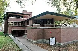 View of the Robie House's front door. There is a concrete path leading from the sidewalk to the front door, which is recessed from the facade. To the right is the living room, which has a brick facade.