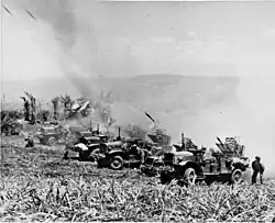 a row of trucks firing rockets. A cloud of dust is behind them.