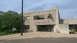 Exterior view of the Smithsonian Institution Museum Support Center, a modern concrete building with brutalist architectural features. The rectangular, multi-story structure has 'SMITHSONIAN INSTITUTION' and 'MUSEUM SUPPORT CENTER' inscribed in capital letters on its tan-colored concrete facade. The building features a central glass entrance with minimal rectangular windows. The foreground shows a paved walkway with a street lamp, while landscaping including trees and shrubs surrounds the building against a partly cloudy sky.