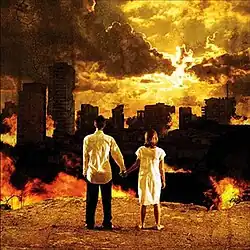 A photo of two people holding hands and facing away from the camera toward a ruined, burning city skyline under a golden sky.