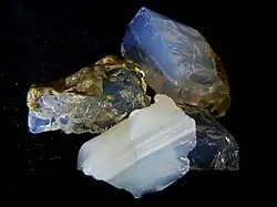 Four opals on a black background. Their opalescence is relatively low, appearing more as translucent blue and white rock than opal proper.