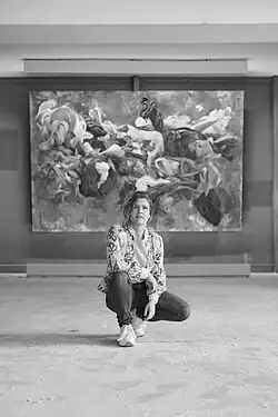 A black-and-white photograph of artist Sacha Lees posing before one of her Baroque-inspired works—a dynamic composition where liberated fabric and entangled limbs collide in chaos.