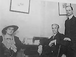 A photograph of a seated woman and man in front of a desk with a standing man behind them and to the right.