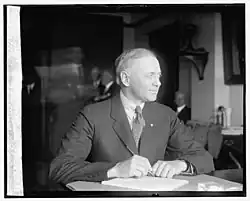 Black and white photograph of a seated, short-haired man looking off to the side and wearing a striped suit and tie.
