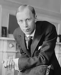 Black and white photo of Prokofiev standing besides a fireplace, his arm rested on the mantlepiece