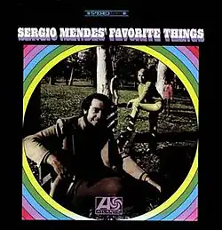 A photo of Sérgio Mendes and a woman resting on a park bench. The photo is surrounded by a circular rainbow.