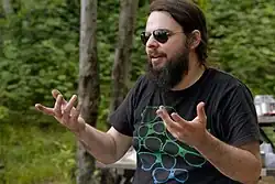 McGrath gesticulates with both hands while looking to the left. He wears aviator sunglasses, a black T-shirt with a sunglasses graphic, and has a long, brown beard. He is in an outdoors, camping setting.