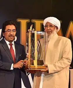 A man in suit and tie hands a large glass and wood award statue to a man in sheikh robes on stage