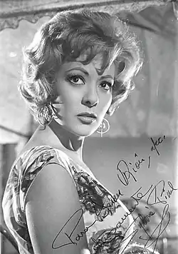 Upper-body shot of Silvia Pinal, a white woman with wavy blonde hair wearing a light dress, looking back at the viewer. The photographer features her autograph on the corner.