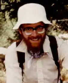 A bearded man in a white shirt and outdoor hat, crouching, with a backpack on his back.