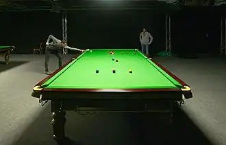 A player taking a shot at a practice snooker table, photographed from the opposite end of the table using a low camera angle to give forced perspective