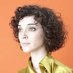 Three-quarter angle profile of St. Vincent wearing a yellow shirt against an orange background