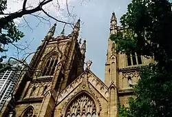 View looking up between Plane trees towards two towers with a gable between them. The towers and gable have panelling to match the window tracery and curvilinear ornamentation.