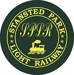 Silhouette of a steam locomotive with cow catcher and smoke plume below the cursive letters "SPLR", surrounded by the words "STANSTED PARK LIGHT RAILWAY" arranged in a circle, all in yellow on dark green