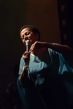 A woman in a blue dress holding a microphone with her right hand and smiling to the ground.