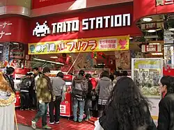 Photograph of numerous people walking by the storefront and into the entrance of a Japanese business. The sign above the entrance reads in English "TAITO STATION" with a pixelated alien character to the left of the words.
