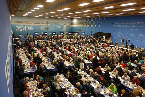 A very large crowd sitting around many tables, playing chess in a large hall