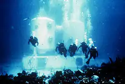 Five scuba divers swim towards the observer in clear water in front of an underwater structure which stands on the seabed in the background, partly obscured by their exhaled bubbles. The white structure comprises two large vertical cylinders on a rectangular horizontal box with access openings.