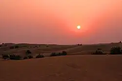 View of dunes at sunset. The setting sun hangs low and dim in a sky graded from fiery orange at top to maroon near the horizon. Terrestrial features are difficult to discern in the crepuscular illumination.