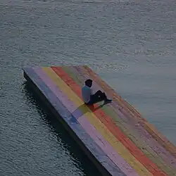 Baby Keem sitting on a rainbow colored wooden pier surrounded by water.