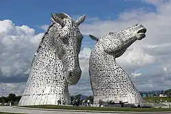 Falkirk High Street with the Falkirk Steeple dominating the centre of the picture. Shops to the left and right are visible with many shoppers on the pedestrianised street.