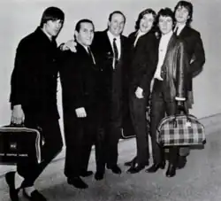 The Kinks and the two executives, all in suits, pose for a group photo, luggage in hand.