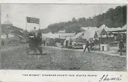 The Midway Richmond County Fair, Staten Island (Concession Stands, people walking about, stadium and Painless Parker's cart in middle of road.