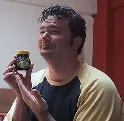Portrait of a smiling man with short dark curly hair. He holds a small jar of Marmite close to his chin while facing left.
